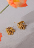 Gold leaf-shaped earrings on a light gray surface with an orange flower in the background.