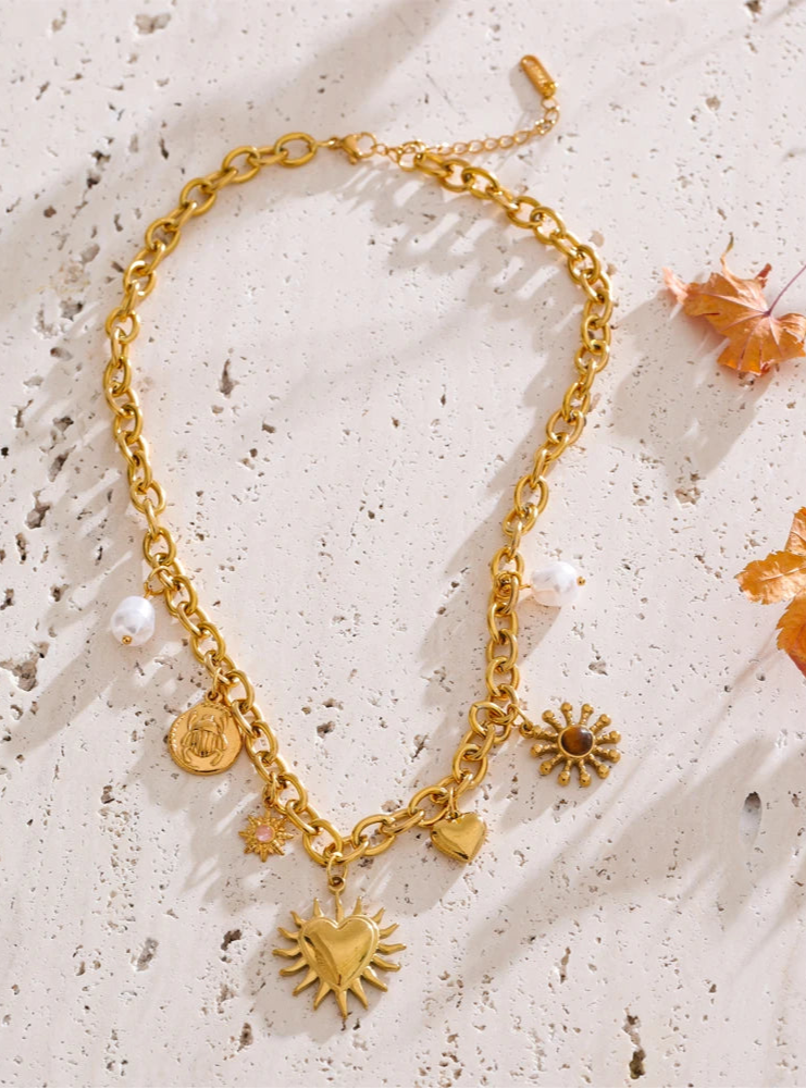 Gold chain necklace with various heart and sun charms on a textured beige surface