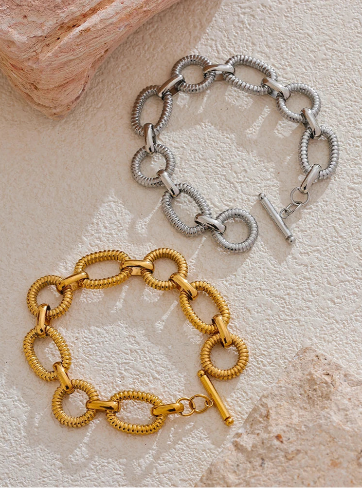 Two chain bracelets, one gold and one silver, on a textured surface.