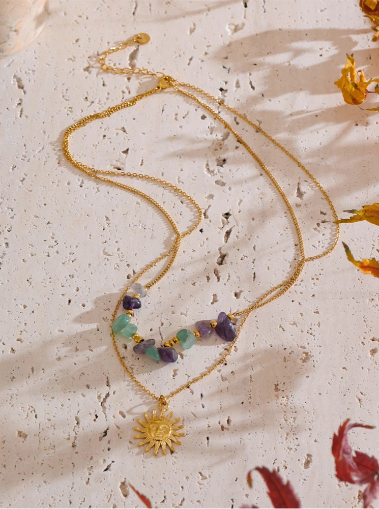 Gold necklace with gemstones and sun charm on a textured beige surface