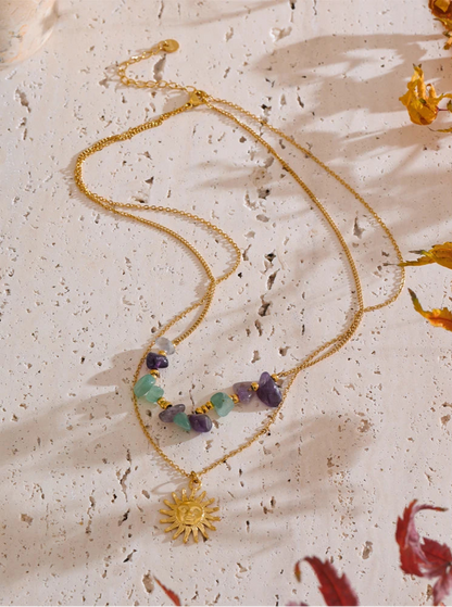 Gold necklace with gemstones and sun charm on a textured beige surface