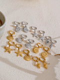 Collection of gold and silver seashell earrings on a textured surface with a cup in the corner.
