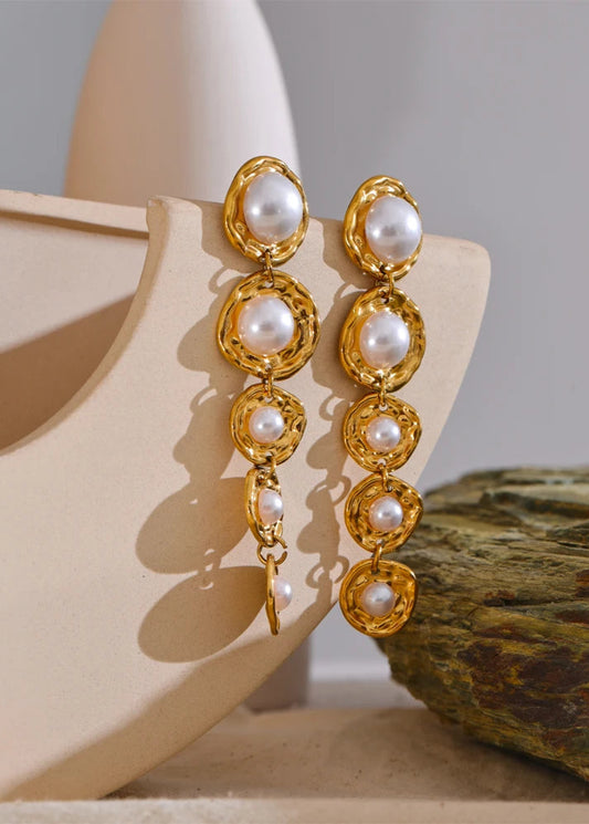 Gold drop earrings with pearl-like stones on a beige stand against a neutral background